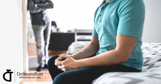 two men in a room, tension, upset, one sitting on bed and one standing in doorway