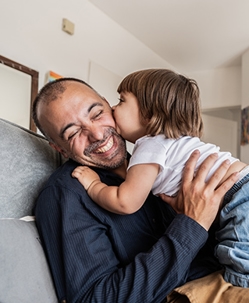 A father working on a laptop in a chair by a window while his daughter sits beside him on the floor doing homework, both focused on their tasks.