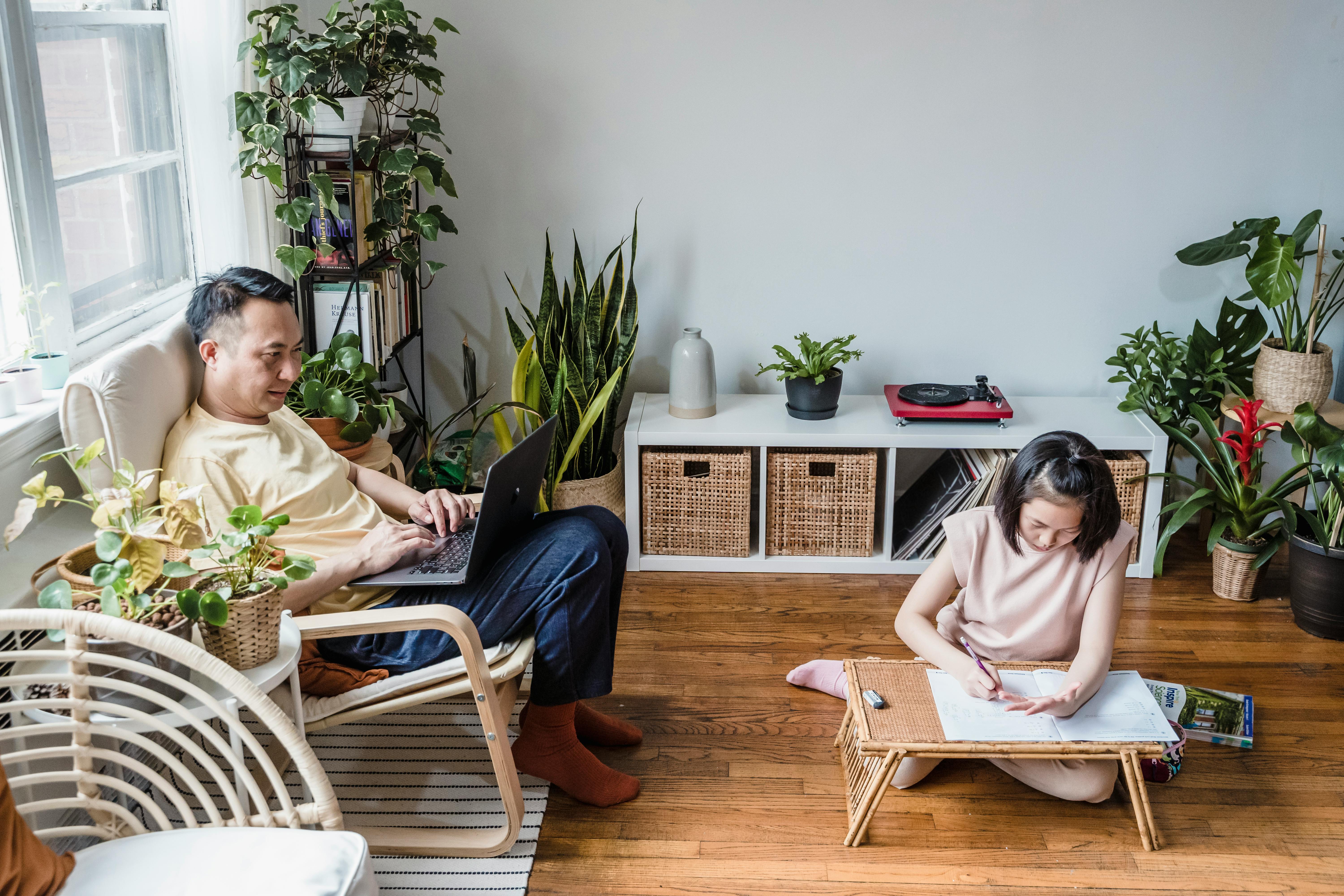 A father working on a laptop in a chair by a window while his daughter sits beside him on the floor doing homework, both focused on their tasks.