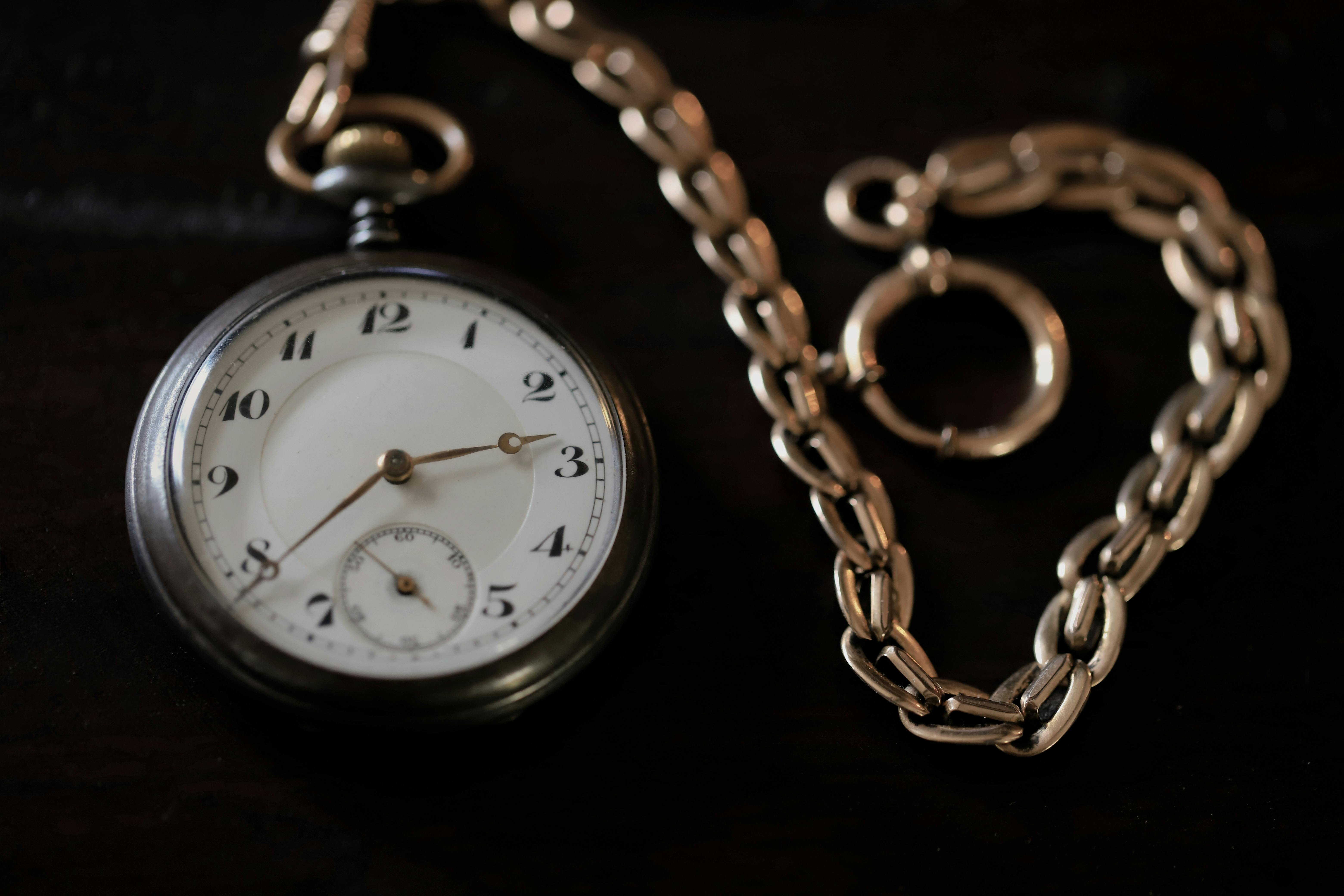 Close-up of a vintage silver pocket watch with a white clock face and black numerals, attached to a gold-toned chain resting on a dark wooden surface.