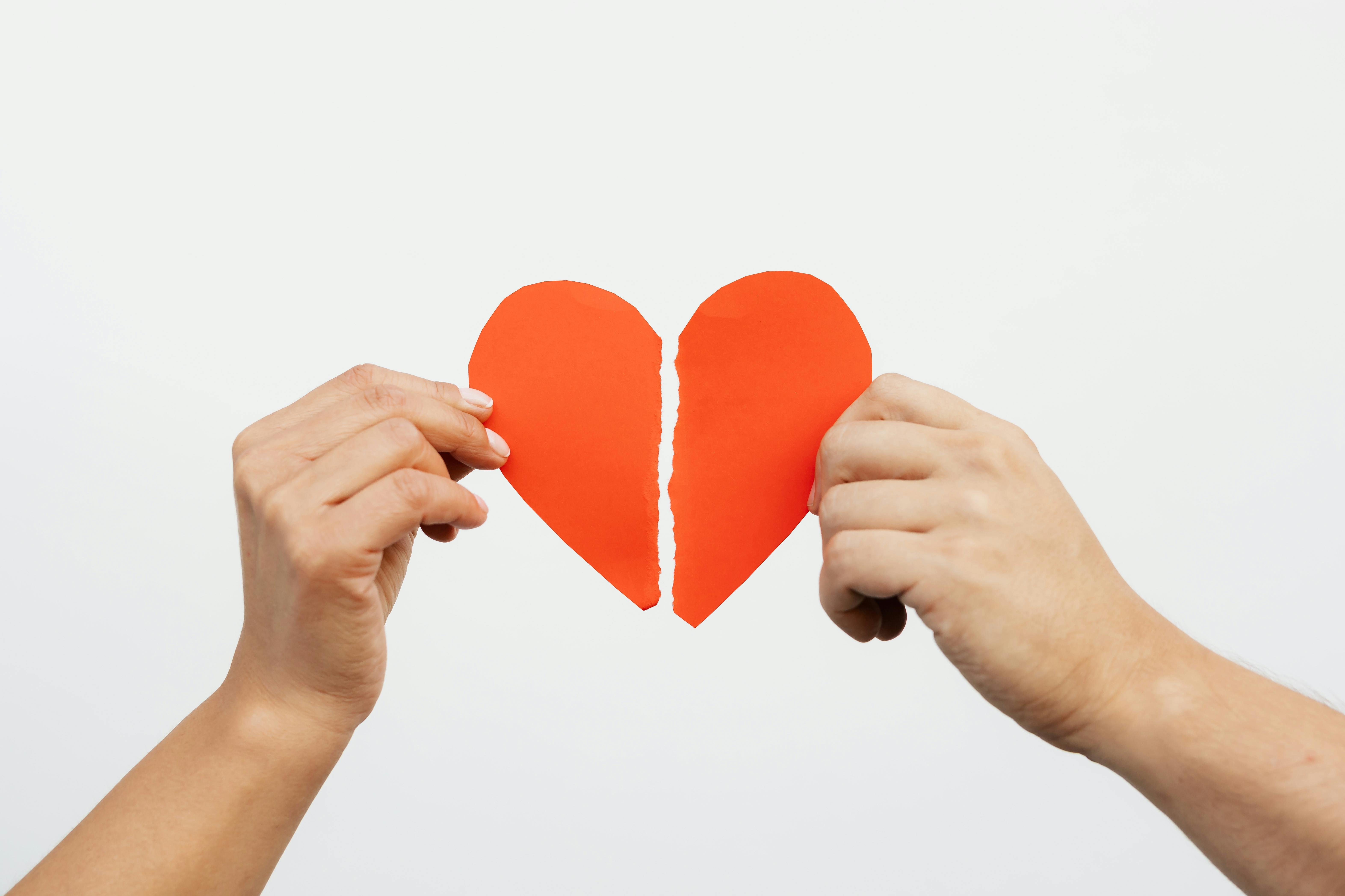 Two hands holding the torn halves of a red paper heart against a plain white background, symbolizing a breakup or emotional separation.