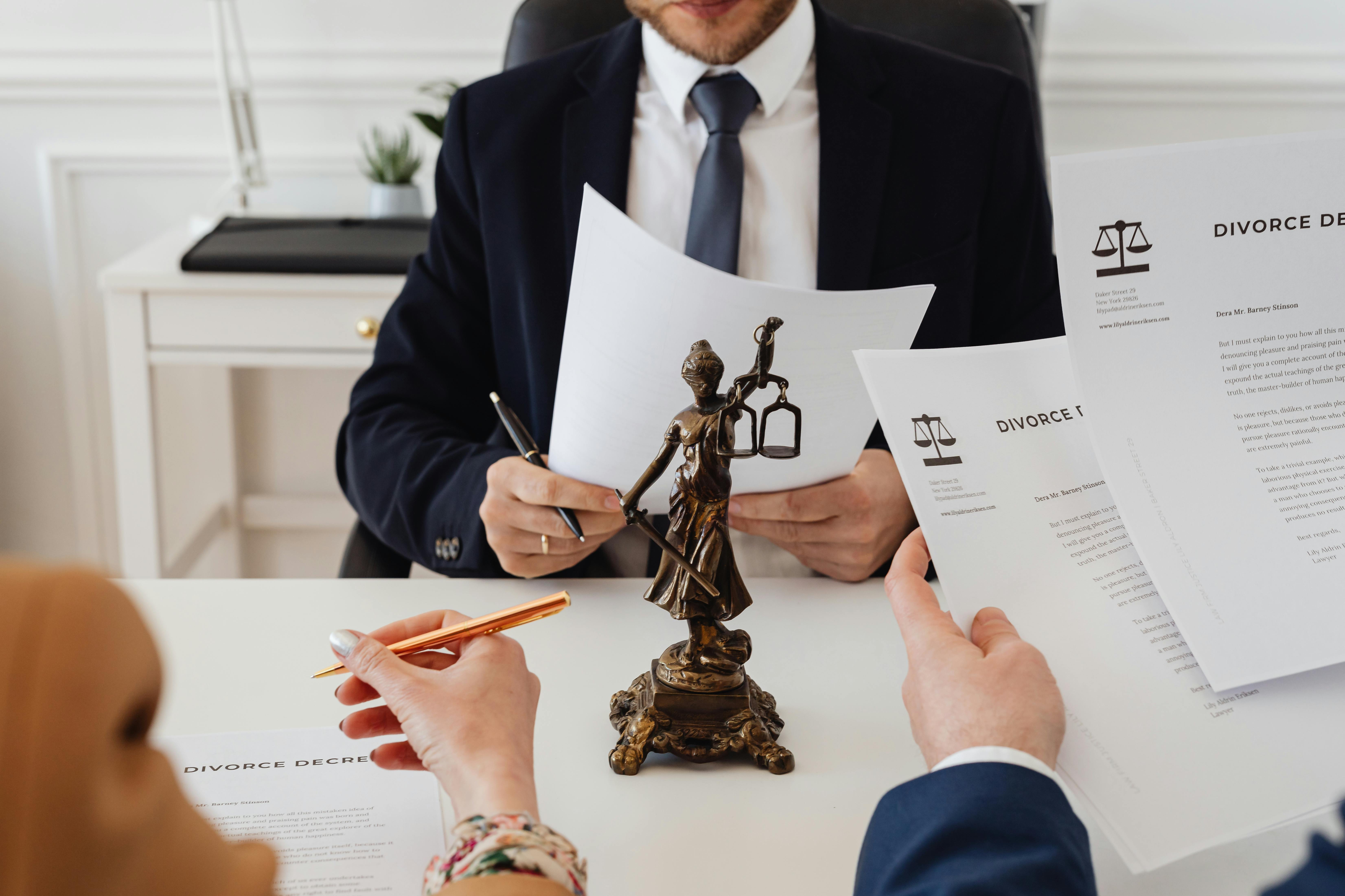 Three people in formal attire reviewing divorce documents at a desk, with a bronze Lady Justice statue centered between them, symbolizing law and fairness during the divorce process.